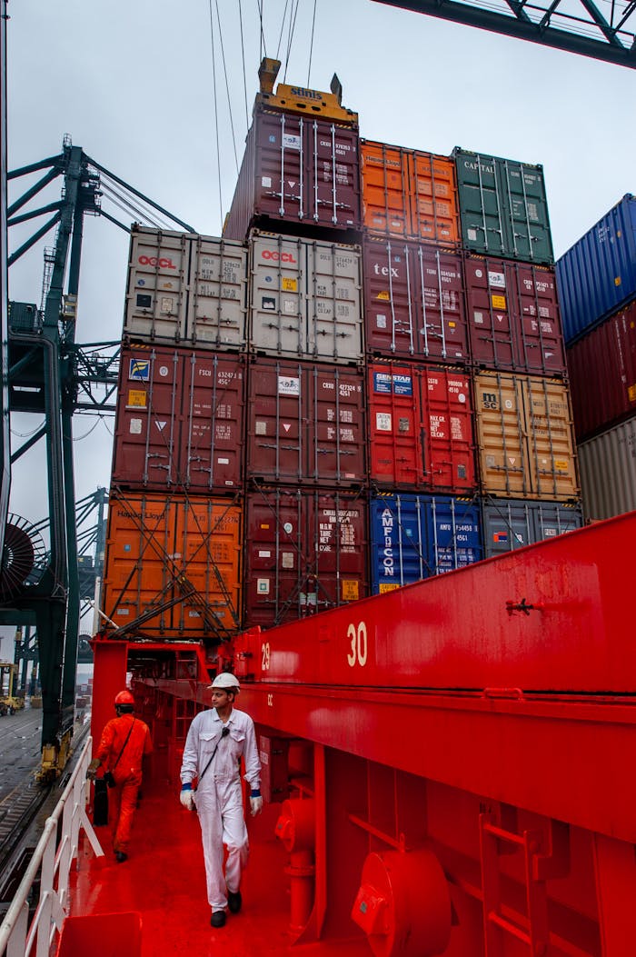 Maritime workers navigating a ship deck with towering stacks of shipping containers in port.